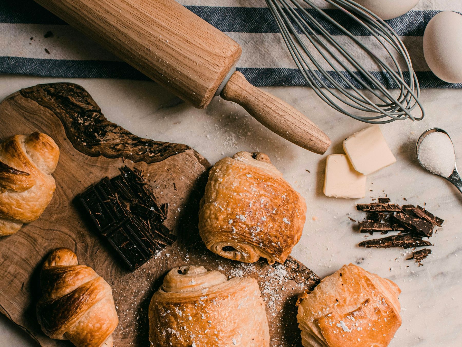 Croissants et pains au chocolat sur une planche, avec du chocolat, du beurre et un rouleau à pâtisserie.